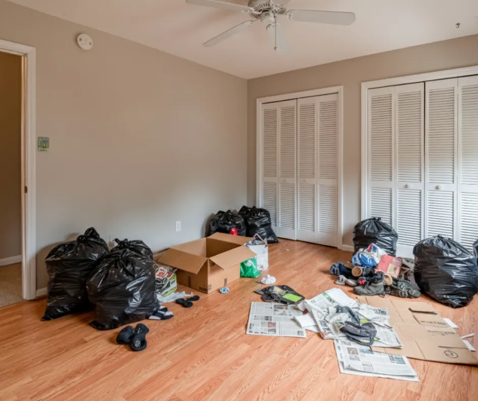Cluttered room filled with trash bags, boxes, newspapers, and junk scattered across hardwood floors
